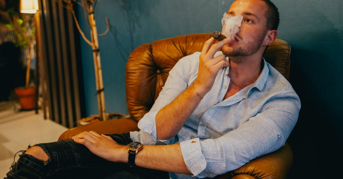 A man in a relaxed pose enjoying a cigar in a cozy armchair indoors.