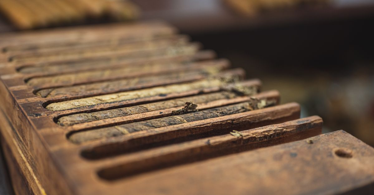 From above closeup of wooden box with narrow rectangular sections with ribbed surface for cigars near table with pile of raw tobacco products in factory