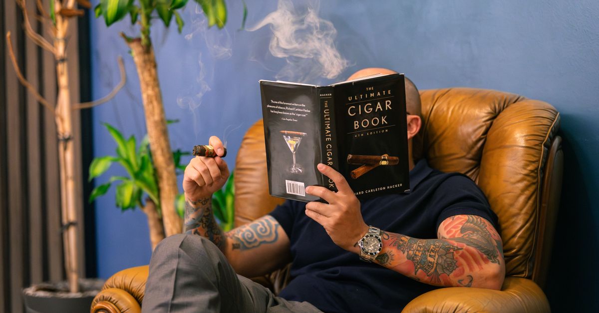 Tattooed man enjoys cigar and book in vintage armchair, creating a relaxed atmosphere indoors.