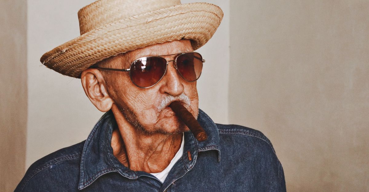 Senior man with sunglasses and straw hat smoking a cigar indoors.