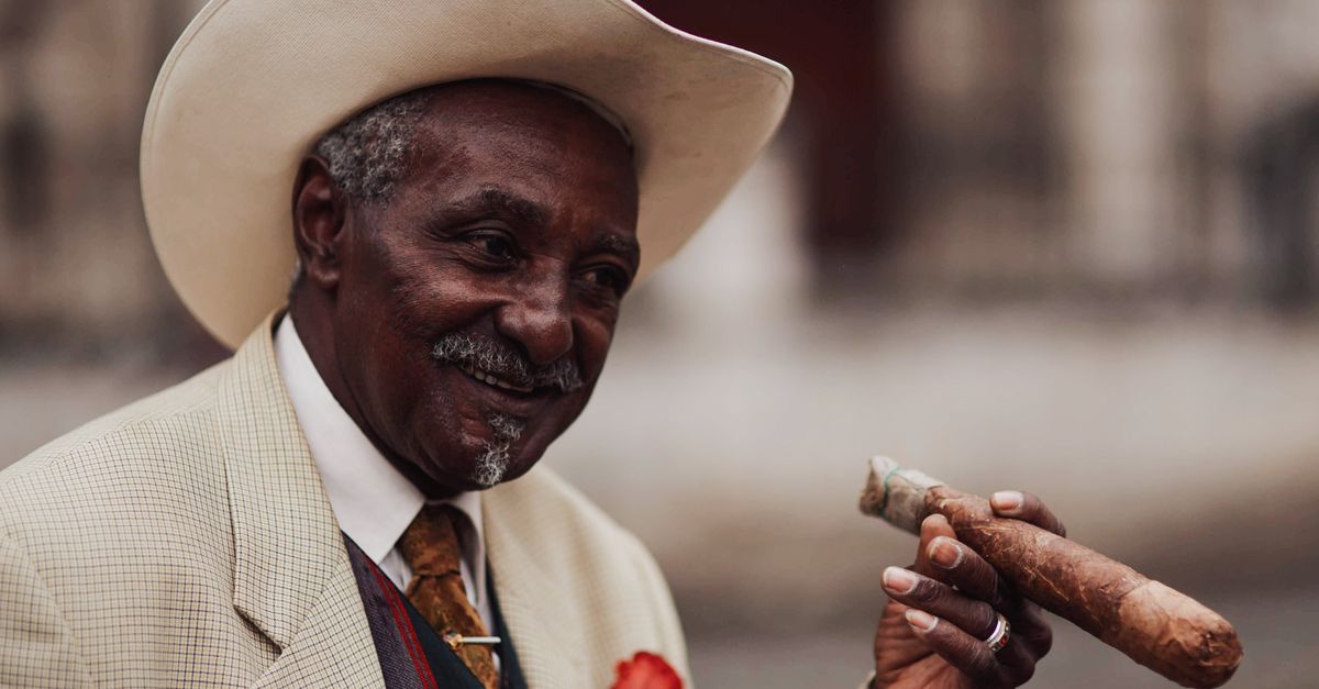 Portrait of a senior man in a cowboy hat and suit, smiling while holding a cigar outdoors.