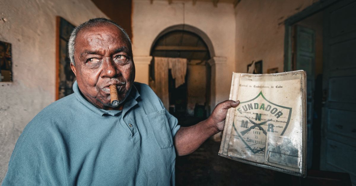 Elderly man smoking a cigar while holding a historical document indoors.