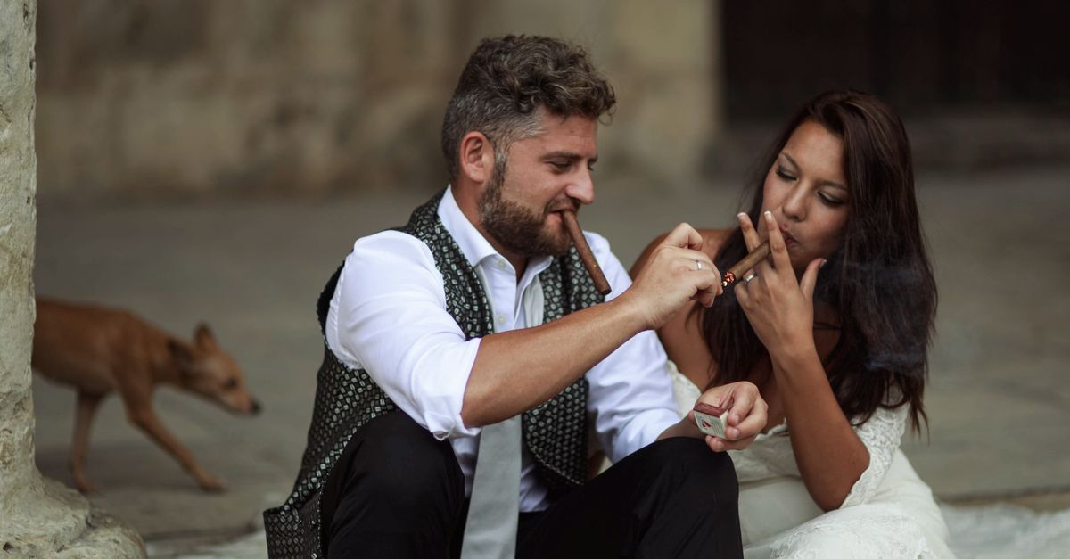 Bride and groom sitting outdoors enjoying cigars together on their wedding day.