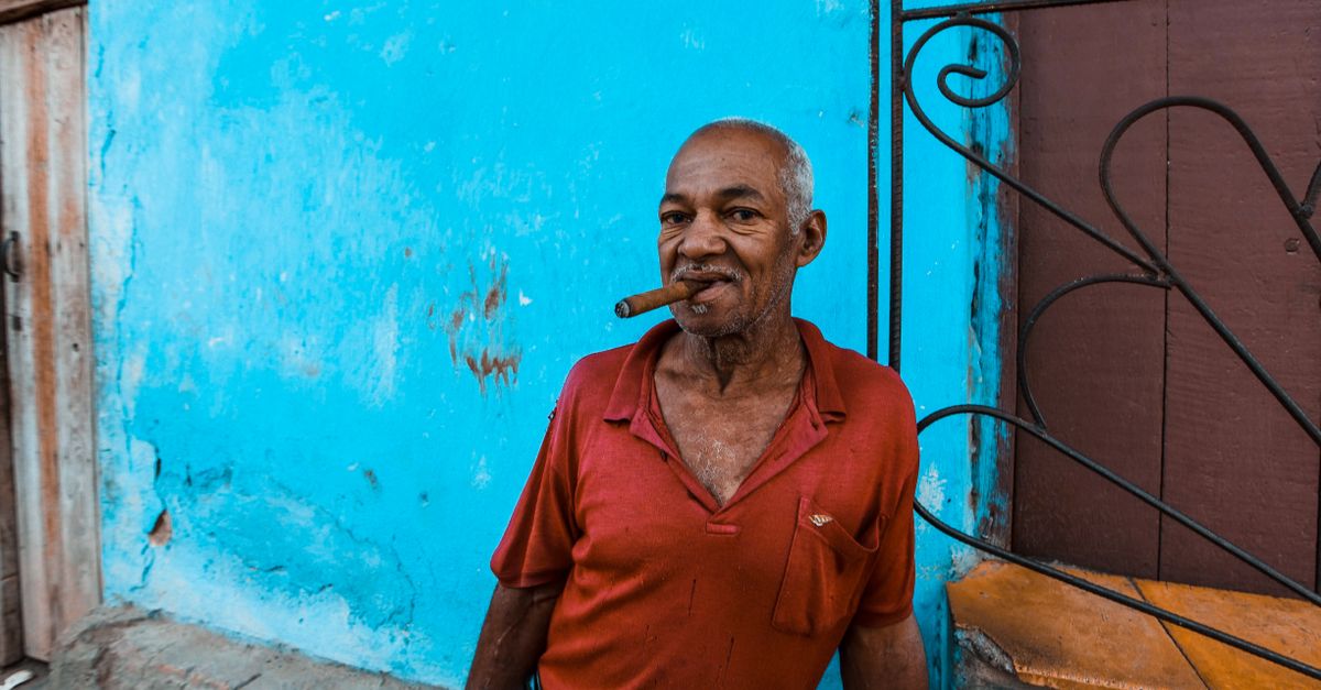 Senior man enjoying a cigar while leaning on a bright blue wall outdoors.